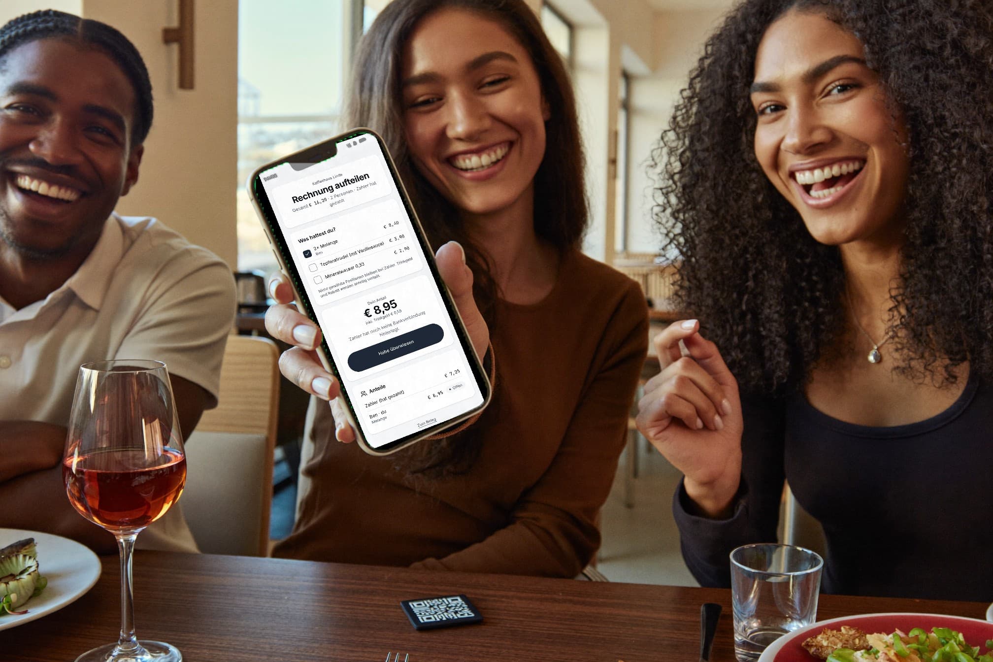 Drei Freundinnen und Freunde lachen am Restauranttisch, eine hält das iPhone in die Kamera: Rechnung aufteilen, Ben hat zwei Melange abgehakt, sein Anteil 8,95 Euro. Auf dem Tisch liegt die kleine Kachel.
