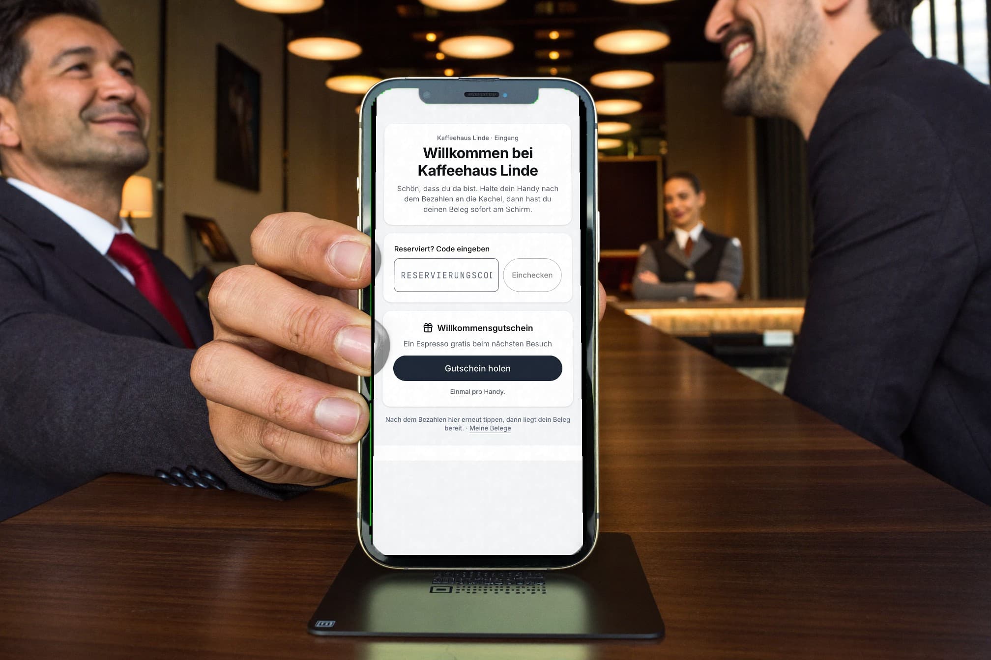 Zwei Gäste an der Hotelbar, einer hält sein iPhone auf der Kachel: Willkommensseite mit Feld für den Reservierungscode und Willkommensgutschein. Die Gastgeberin wartet im Hintergrund.