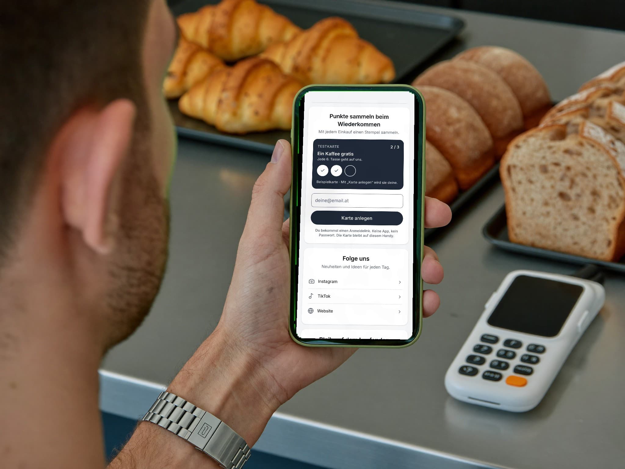 Über die Schulter in einer Bäckerei: Ein Kunde hält sein iPhone mit der Stempelkarte, zwei von drei Stempeln gesammelt, daneben Croissants, Brot und das Kartenterminal.