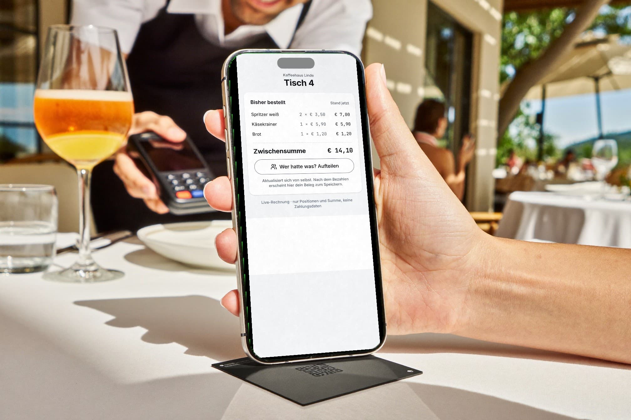 Sonnige Restaurant-Terrasse: Ein Gast hebt das iPhone von der schwarzen Kachel am Tisch, am Schirm die Live-Rechnung für Tisch 4 mit Spritzer, Käsekrainer und Brot. Der Kellner mit Kartenterminal lächelt im Hintergrund.